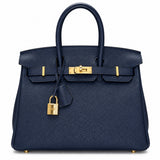 A navy blue pebbled leather handbag with gold-tone hardware, featuring dual top handles, a front strap closure, and a hanging padlock, shown against a plain white background.
