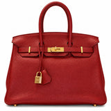 A red grained leather handbag with gold-tone hardware, featuring dual top handles, a front strap closure, and a hanging clochette lock, photographed against a plain white background.