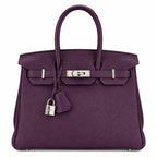 A purple pebbled leather handbag with silver-tone hardware, featuring dual top handles, a front strap closure, and a hanging padlock with clochette, photographed on a plain white background.
