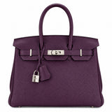 A purple pebbled leather handbag with silver-tone hardware, featuring dual top handles, a front strap closure, and a hanging padlock with clochette, photographed on a plain white background.
