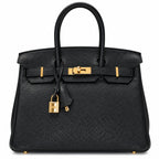 A black pebbled leather handbag with gold-tone hardware, featuring dual top handles, a front strap closure, and a hanging padlock clochette, photographed against a plain white background.
