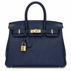 A navy blue pebbled leather handbag with gold-tone hardware, featuring dual top handles, a front strap closure, and a hanging padlock, shown against a plain white background.
