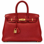 A red grained leather handbag with gold-tone hardware, featuring dual top handles, a front strap closure, and a hanging clochette lock, photographed against a plain white background.